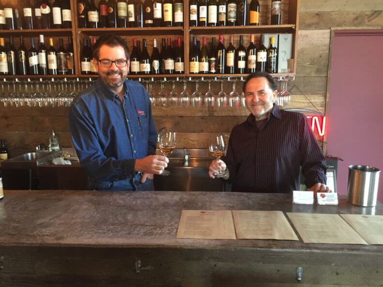 Two men standing at the Wine Country Connection tasting bar with wine bottles displayed behind them